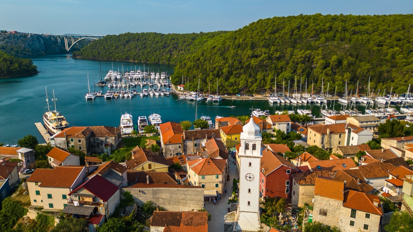 Luftaufnahme der roten Dächer und des Glockenturms von Skradin über dem Jachthafen, dahinter eine Bogenbrücke über den Fluss